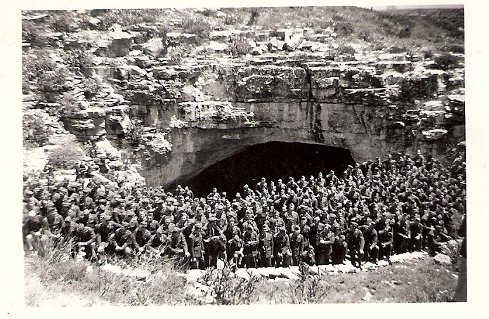 Very large group of men in uniform standing in front of large cave opening.