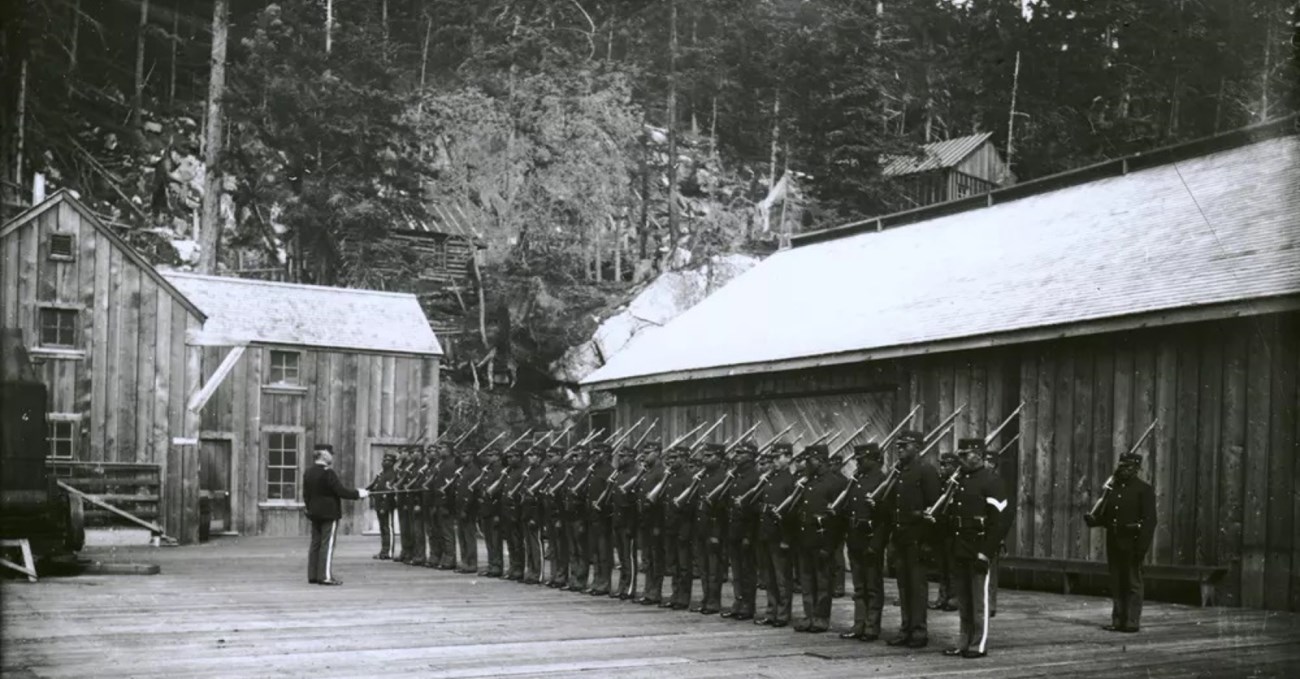 Black and white photo of american american men in army uniforms standing shoulder to shoulder in two rows in formation. They are all standing at attention. Their officer is standing in front of them.