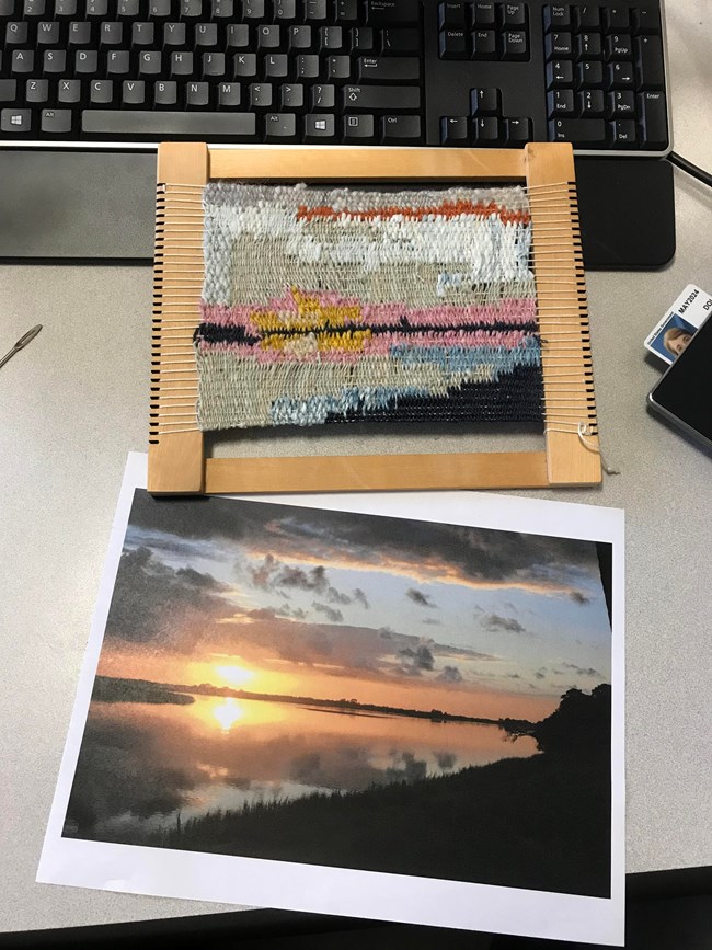 a photo of sunrise on a river sits on a desk next to a small loom with yarn that looks like the picture