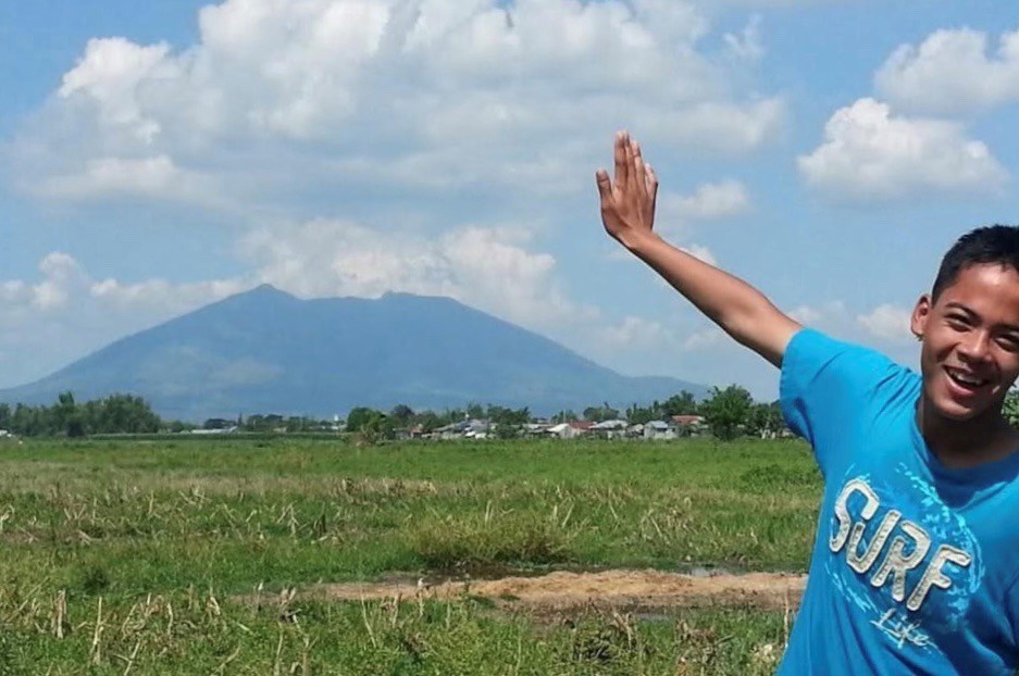 The author as a teenager wearing a blue "Surf Life" t-shirt, posing in an expansive grassy field with his arm extending up over a distant mountain with a distinct volcanic profile.