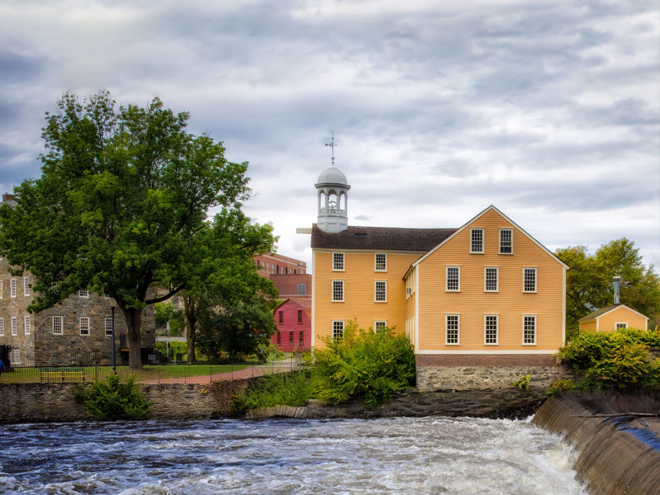 Cloudy day view looking across the river at a yellow three-story building (center right). On the left is a multi-story stone buildingpartly obscured by a large tree, and in the back center, there's a bright red two-story house.