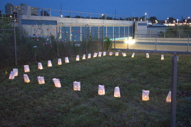 Glowing white paper bags standing side by side, forming a large rectangle on an empty grass patch next to a street.