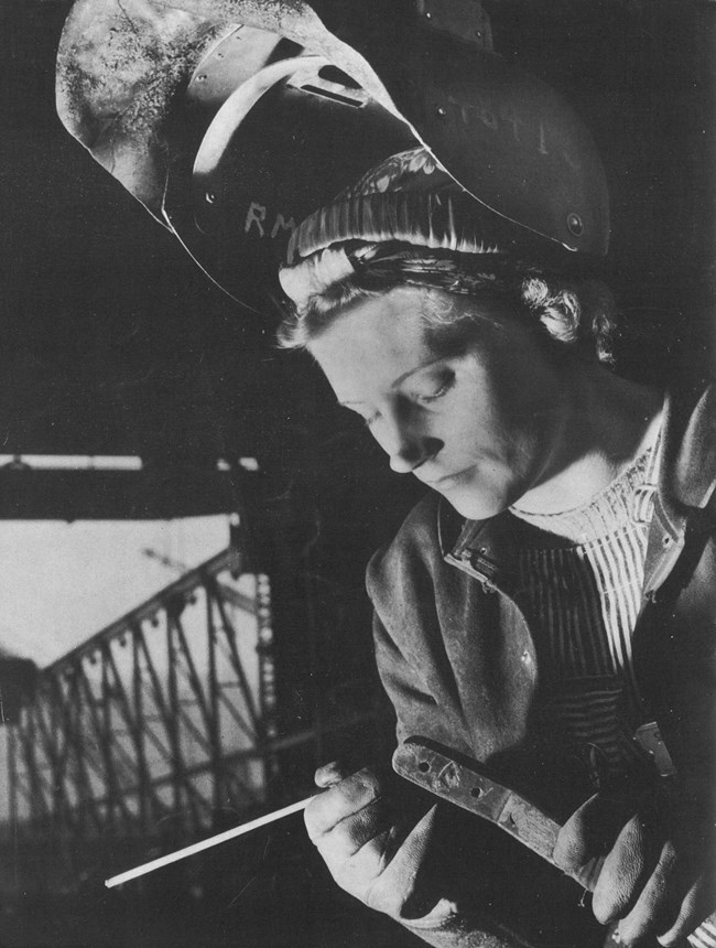 An image of a woman wearing a welding helmet and gloves works on a welding project.