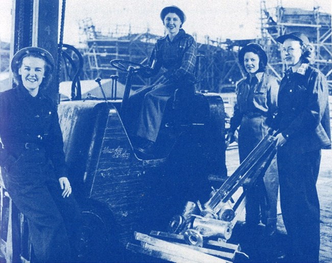 An image of four women working together in a shipyard. One woman sits at a shipyard control panel. Another woman holds a piece of shipyard equipment. They are wearing safety apparel developed for women to wear at the shipyards.