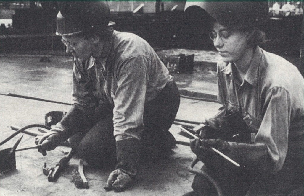 An image of two women sitting together on a ship floor, wearing helmets and other safety equipment, welding pieces of metal together