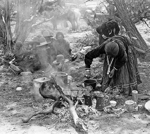 Women bend over harvesting and cooking pinon nuts while there is a crowd of people sitting on the ground in the left background.