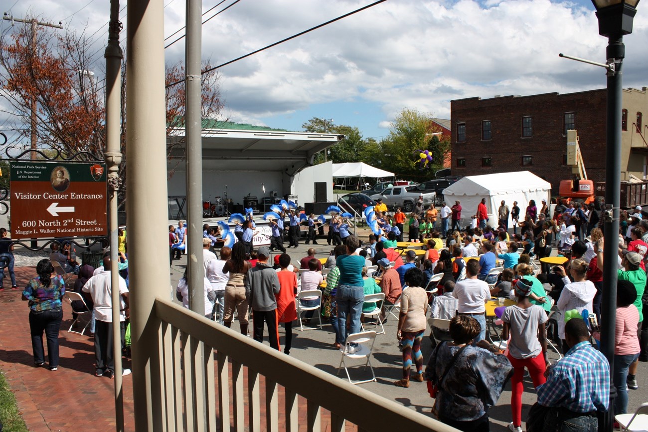 Large crowd gathered on a paved area in front of a stage, viewed from the porch of Maggie L. Walker National Historic Site, with a visitor center sign visible in the far left of the frame.