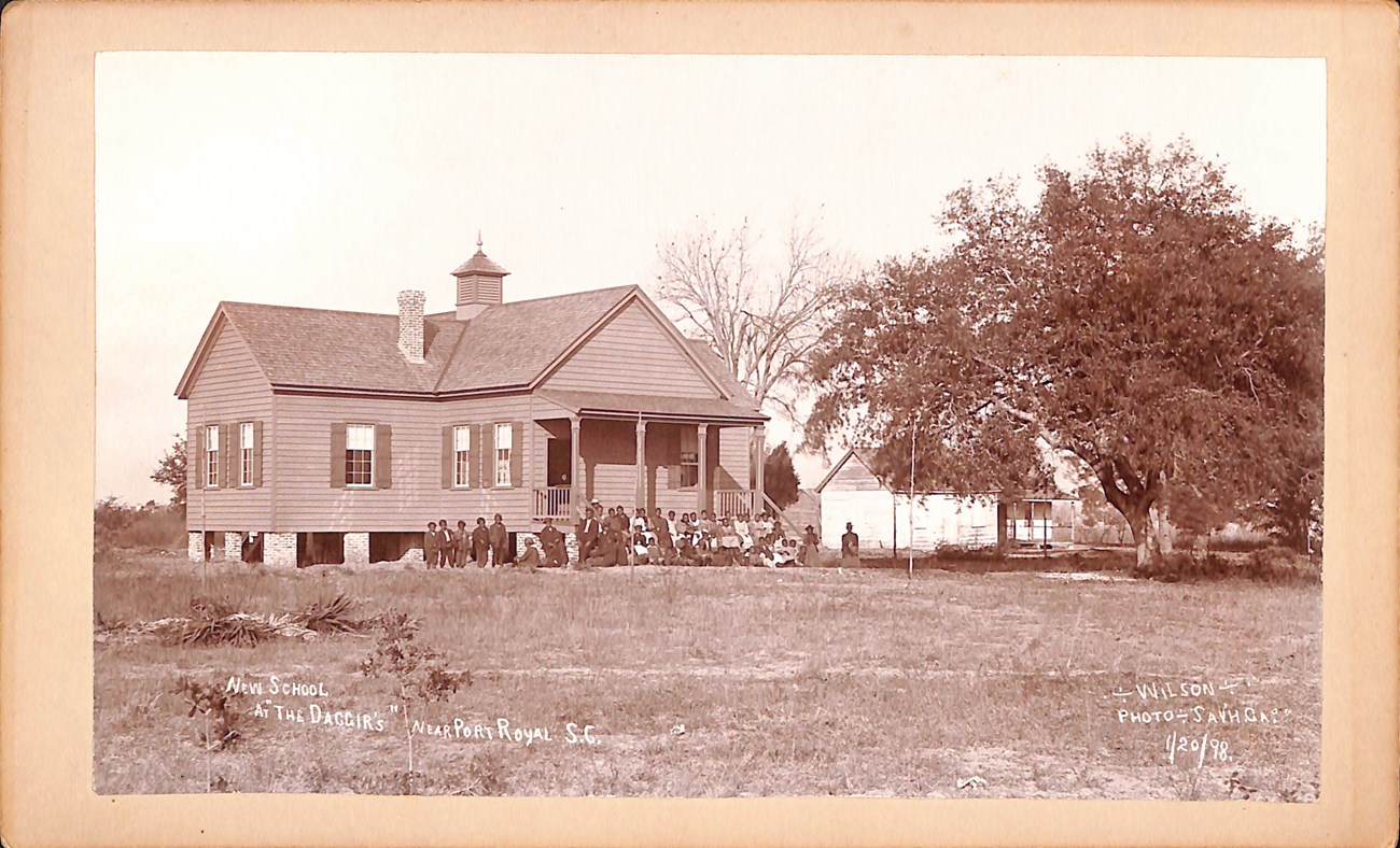 A small wooden school building with children sitting and standing on the front steps.