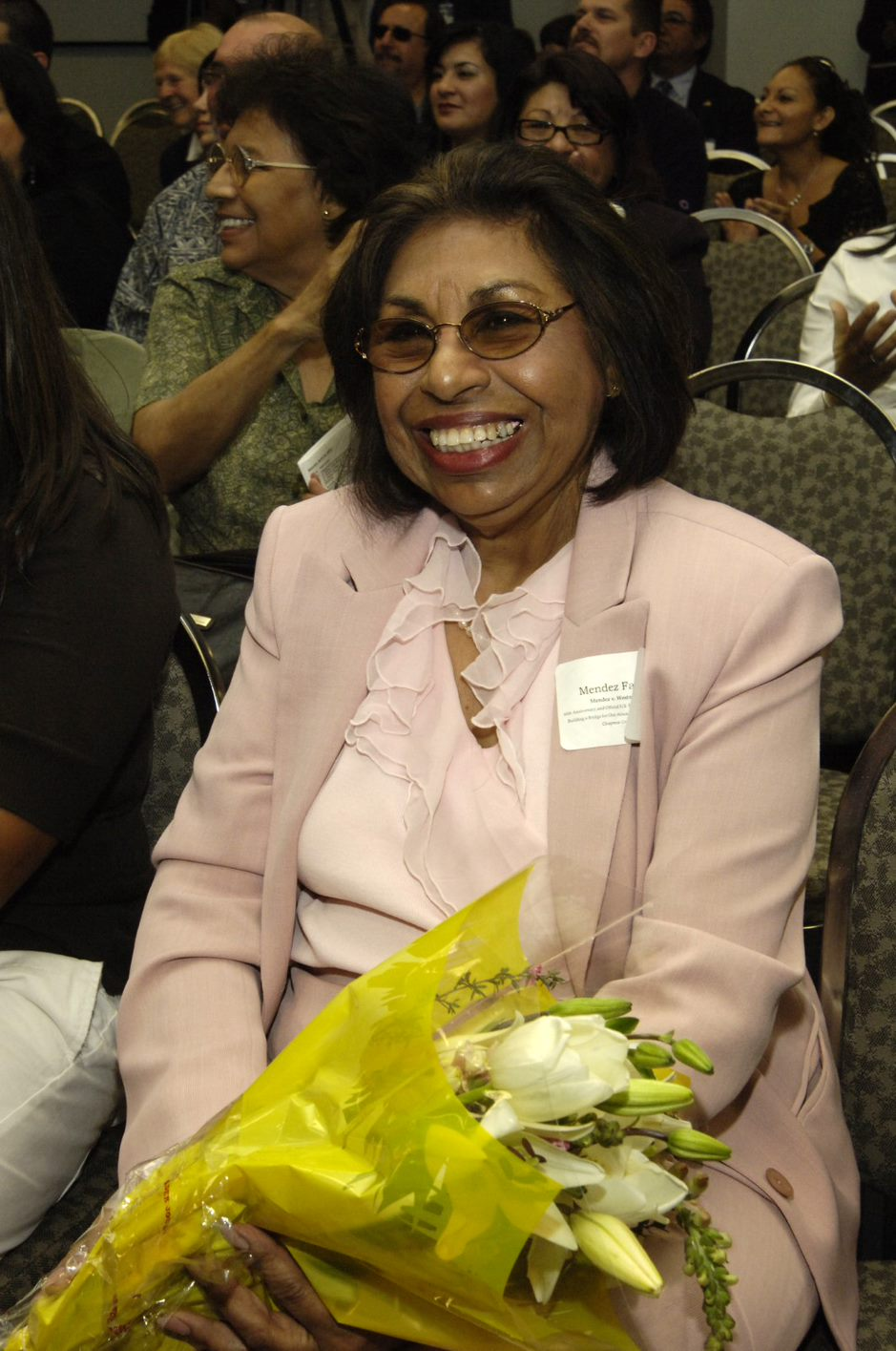 Color photo of Sylvia Mendez as an adult wearing a pale pink skirt suit and blouse and lightly tinted oval glasses. She is seated with a bouquet of flowers and smiles toward the camera.