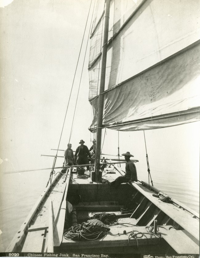 Three people in hats sit aboard a fishing boat with sails.