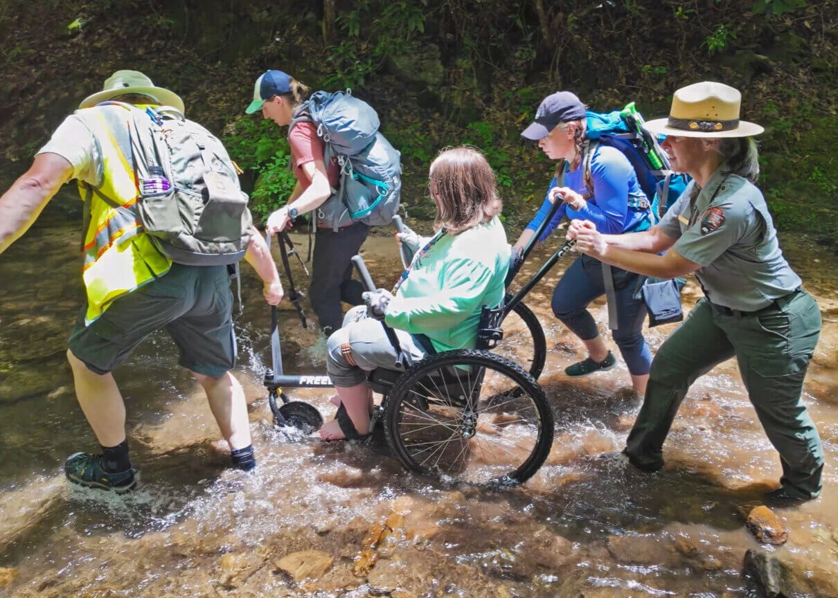 Four people help someone in an off-road wheelchair cross a shallow and rocky stream.