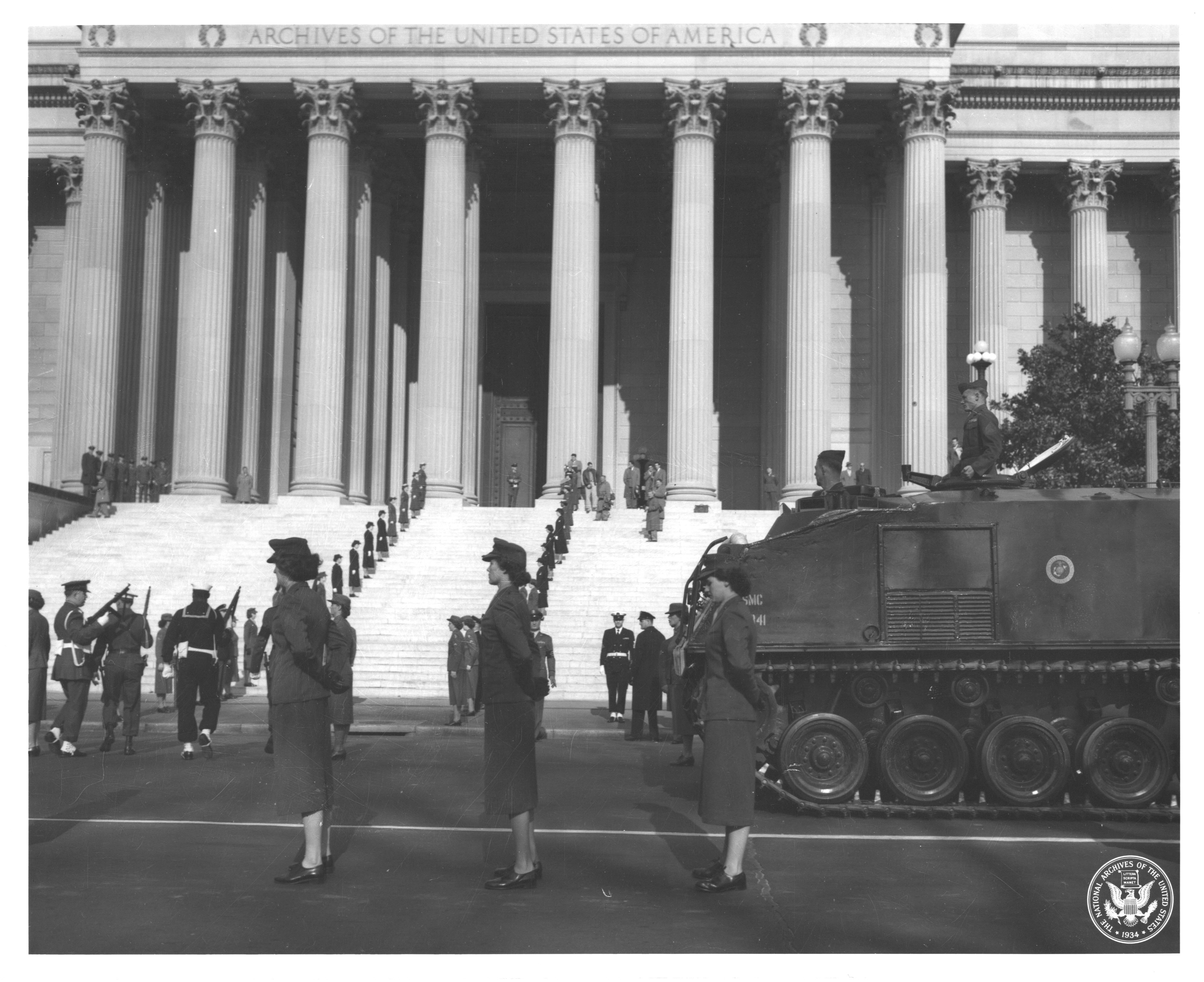 A procession of military personnel, both men and women, with an armored tank in front of the National Archives building.