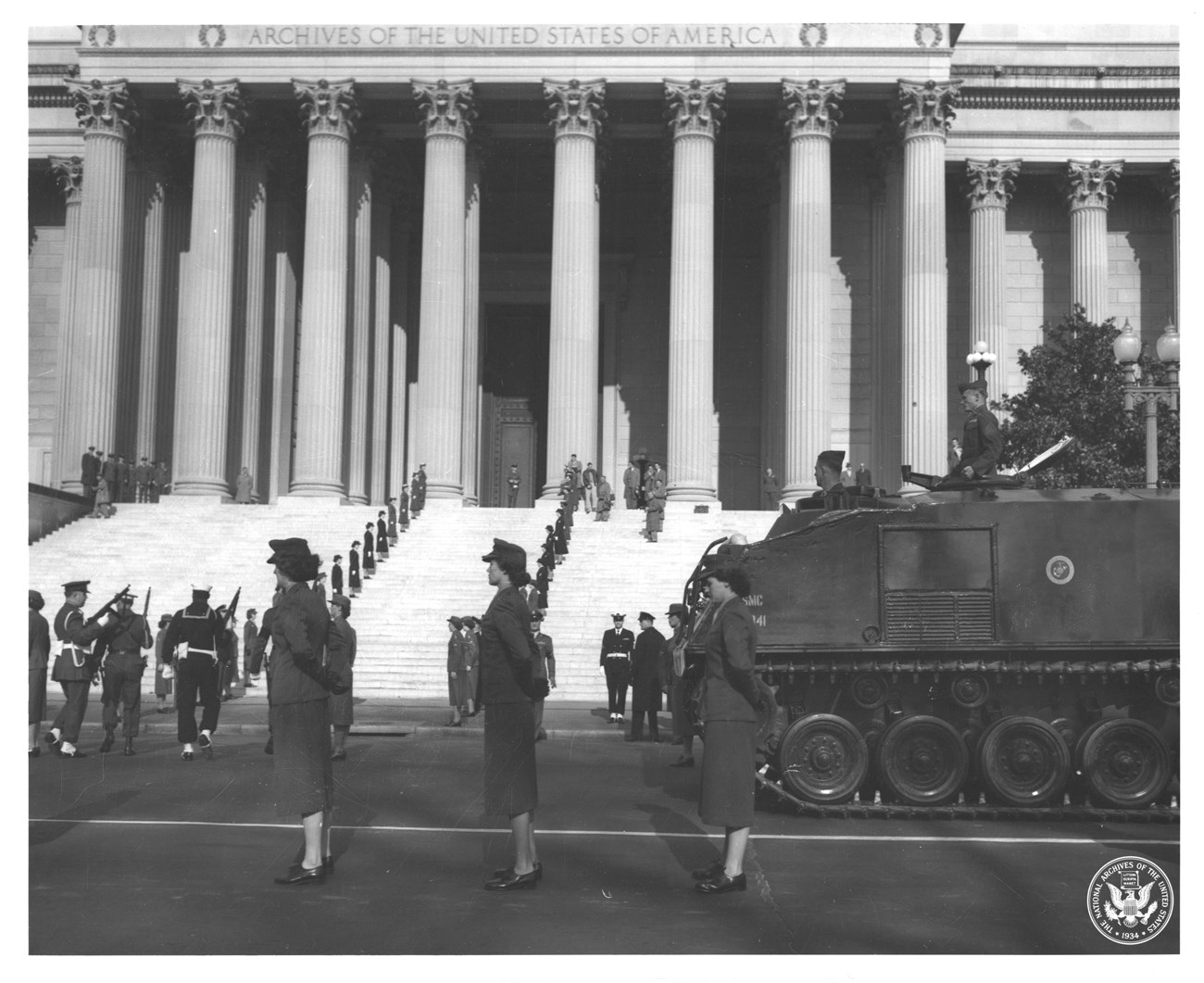 A procession of military personnel, both men and women, with an armored tank in front of the National Archives building.