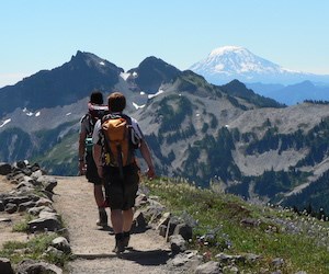 Two hikers with day packs walk along a trail with views of mountain ridges and a distant volcanic peak.