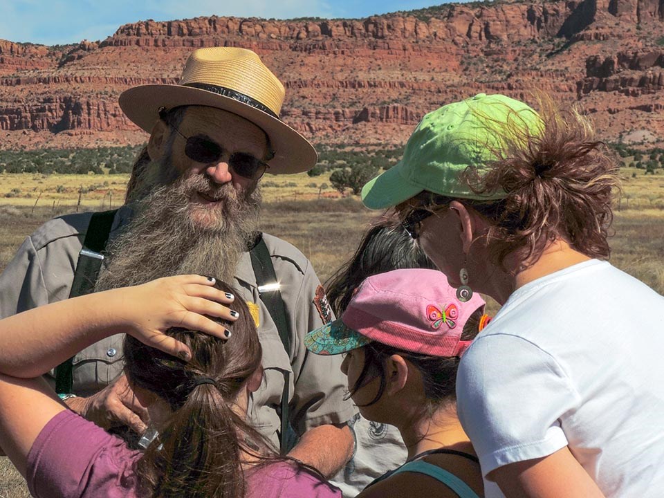 Park Ranger with a flat hat, dark glasses, and a long beard talks to an adult and children while tall red cliffs tower in the background.