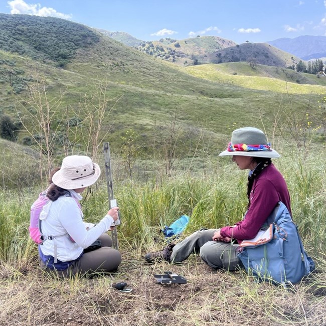 Two adults sit on the grass in front of a mountainous background, holding materials to set up a camera.