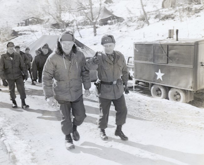 A black and white image showing Dwight Eisenhower walking alongside Major General James C. Fry in a wintry scene in Korea, with each wearing heavy winter coats