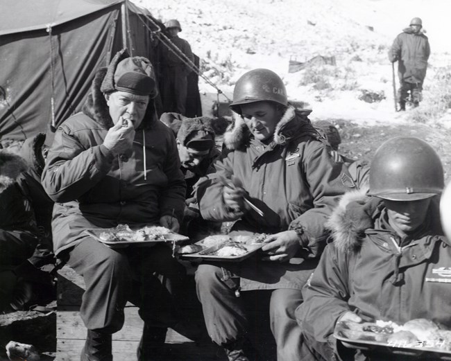 A black and white image showing General Dwight Eisenhower eating with soldiers outdoors in Korea