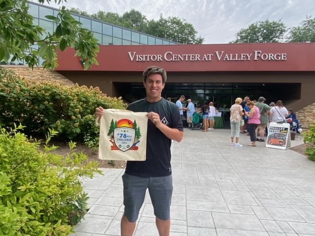 A smiling man standing in front of the Valley Forge visitor center holds up a tote bag with the 2024 logo on it