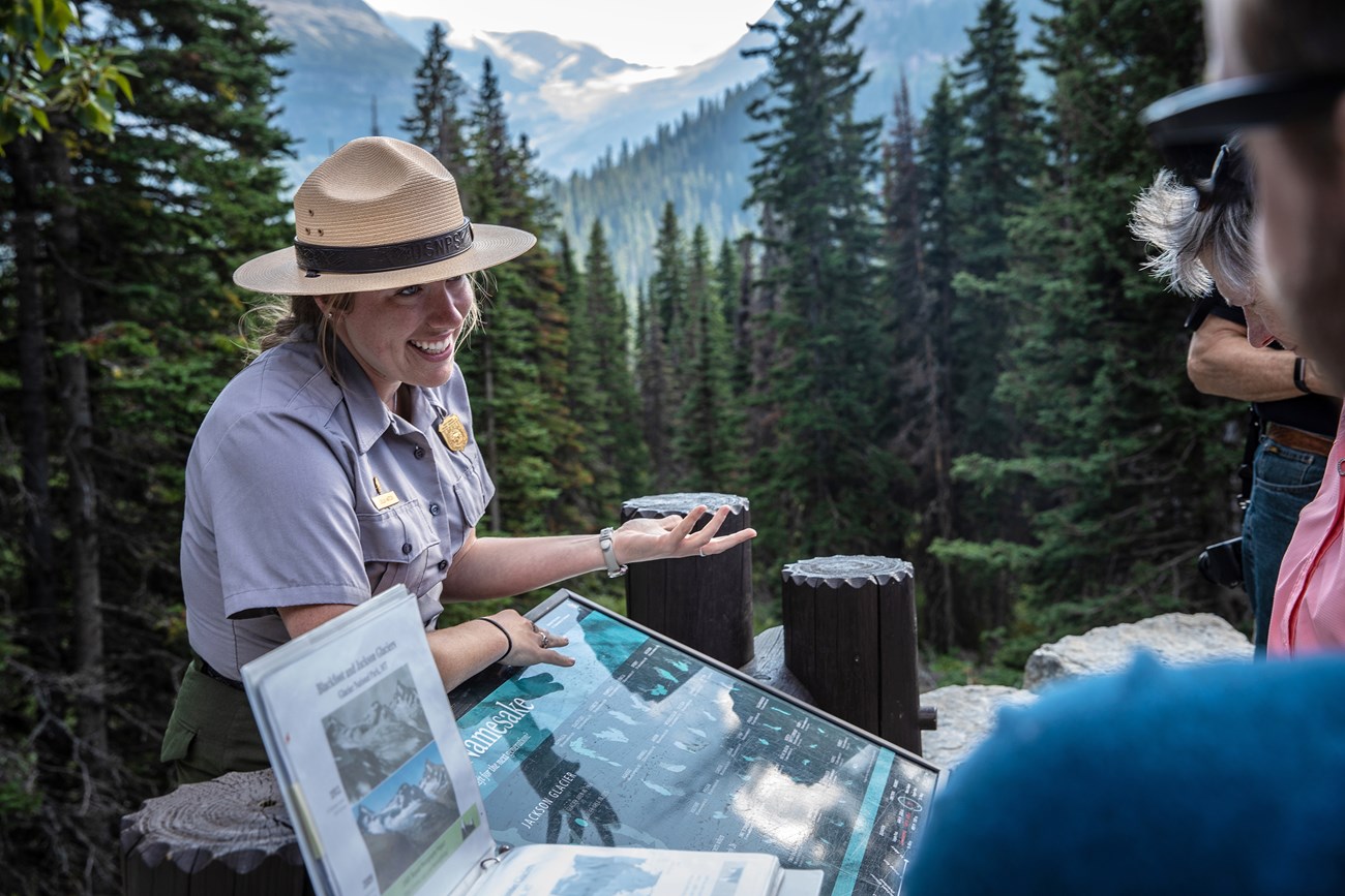 A park ranger leans on a sign while talking with visitors against a backdrop of woods and snow-capped mountains