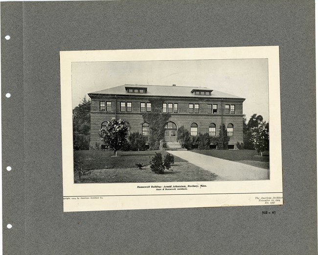 Black and white photograph of stone square building with some vines creeping up the sides. In front of a grassy area with some small trees on it.