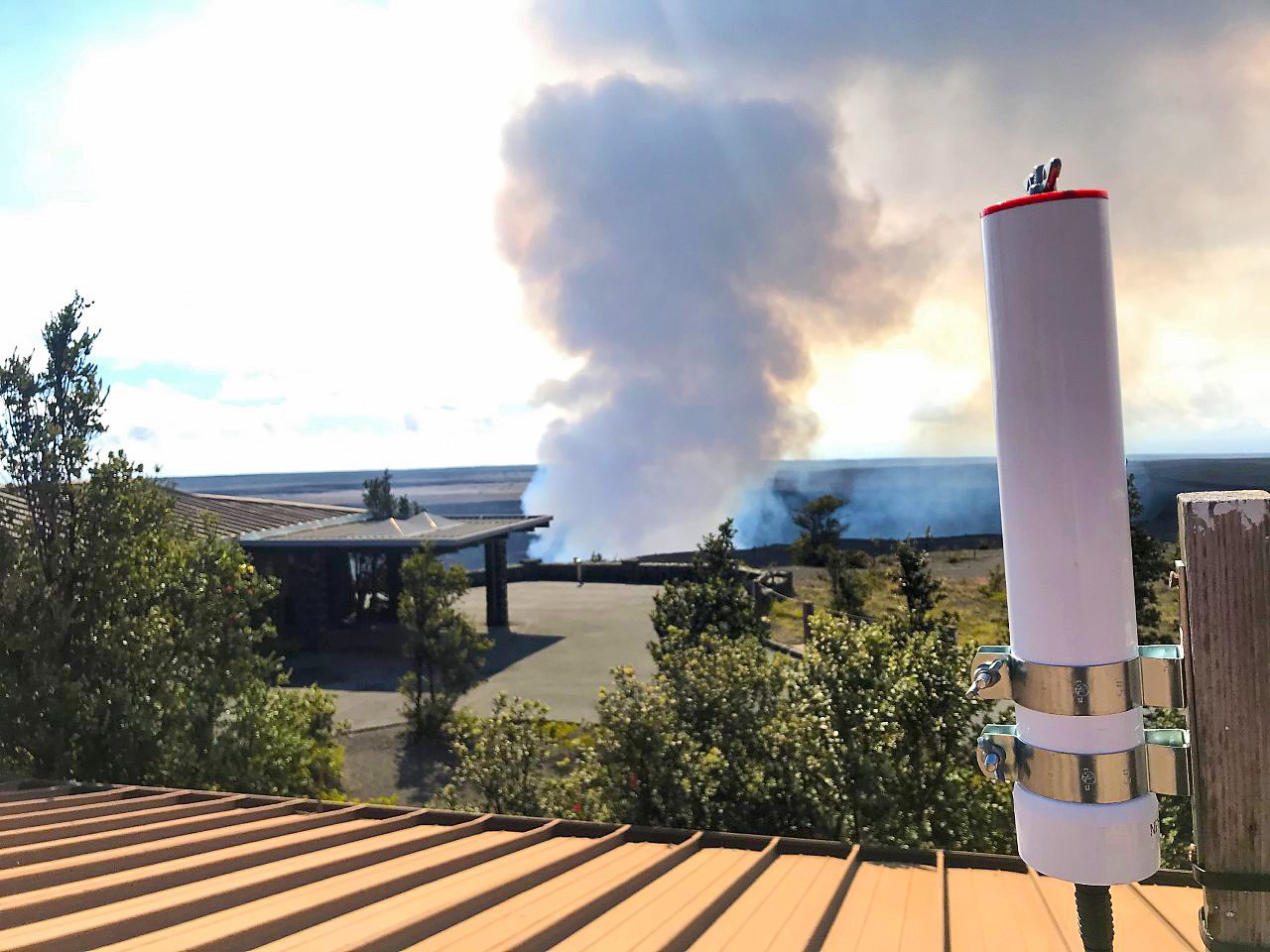 White cylyndrical device mounted to a post on a rooftop overlooking Kīlauea Volcano as it spews a plume of gasses into the air.