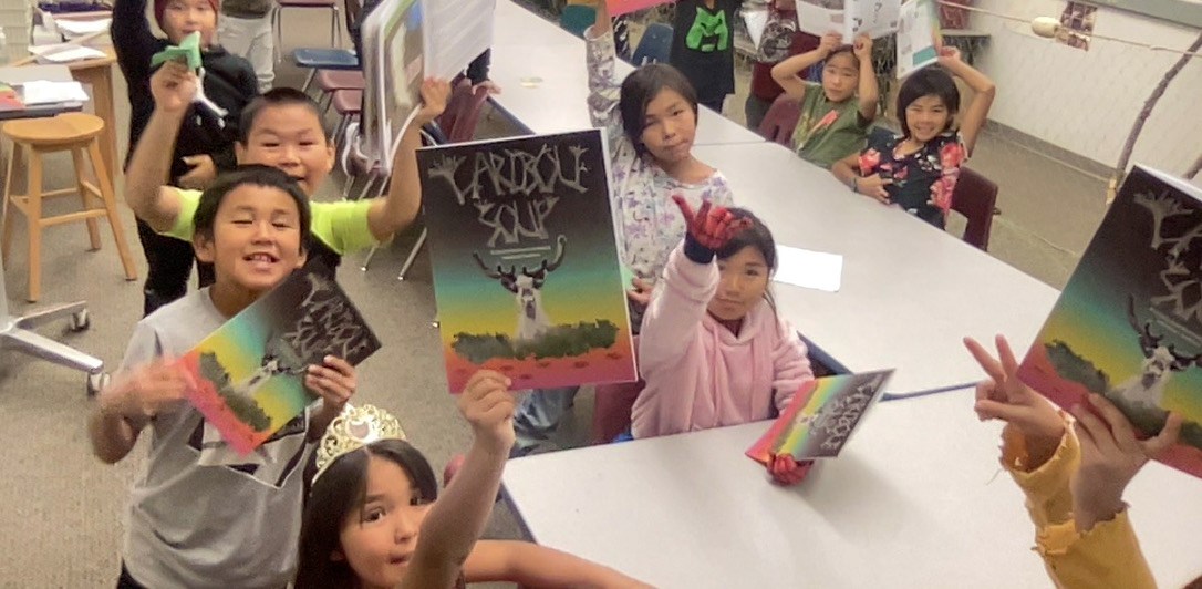 Young students hold up workbooks titled "Caribou Soup" in a classroom.