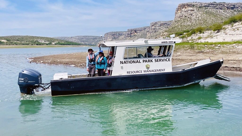 Four park staff members on a boat next to the shore of a reservoir surrounded by desert hills. The boat has writing that says, "National Park Service Resource Management."