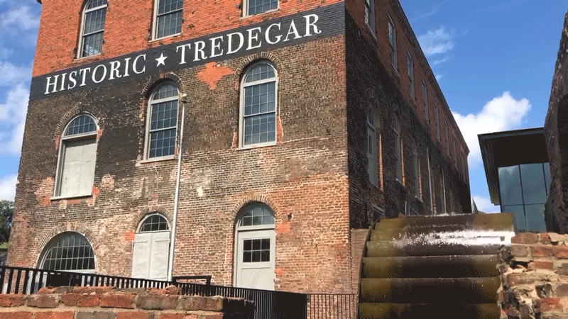 Water wheel spinning, with water splashing and glistening over it in the sun, next to a three-story brick building with the text "HISTORIC TREGEDAR" painted on the front of it just above the second story windows.