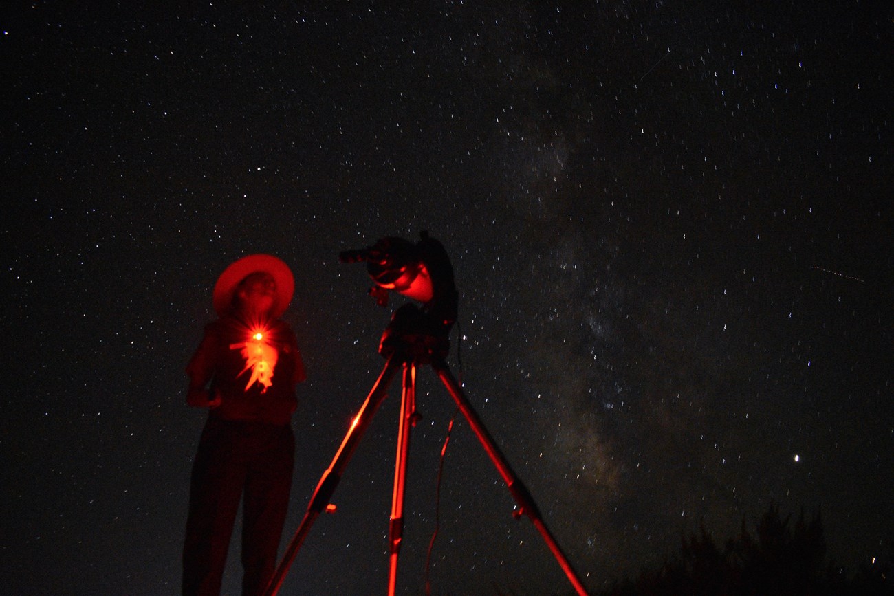 a ranger and a telescope illuminated in red with a starry sky overhead