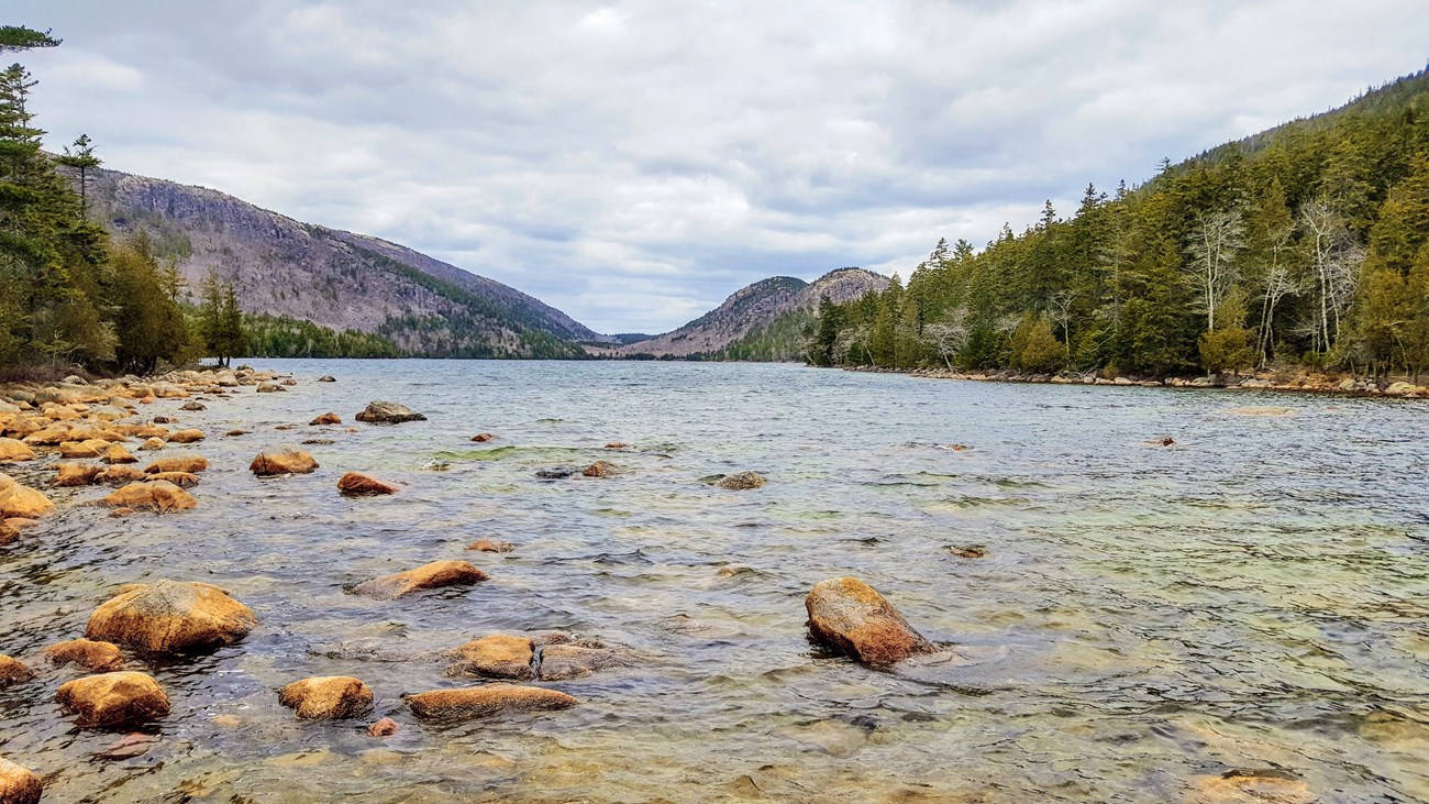 Scenic lake on an overcast day, backed by rocky and tree-covered, rounded mountains. Small boulders line the shore and are scattered throughout the shallower waters of the foreground.