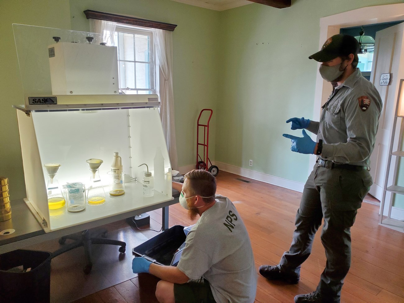 Two men with masks and NPS uniforms look at chemicals in glassware stored inside a fume hood.