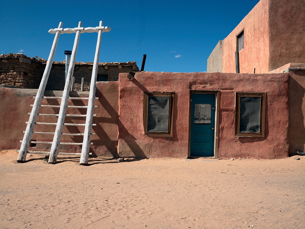 Single story plaster building with a blue door inbetween two windows and a ladder to the roof on the left