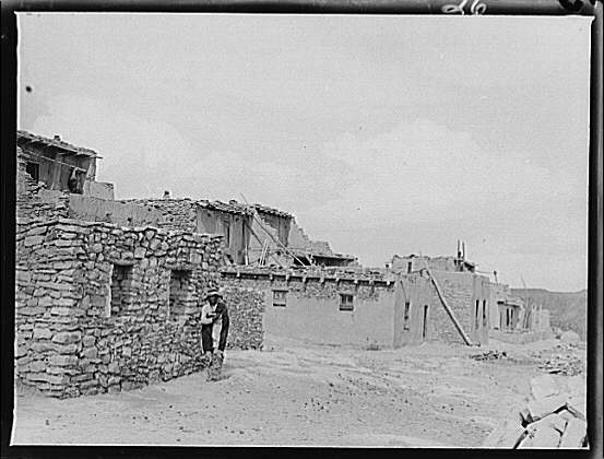 Black and white image of a few brick buildings in Acoma Pueblo with a man standing next to one for scale