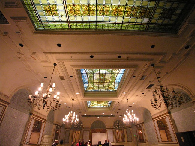 Ornate white ceiling featuring green, blue, and brown stained glass skylights and chandeliers and arched alcoves forming upper third of side and back walls