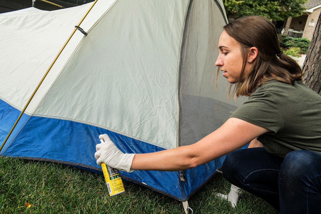 A woman wearing disposable gloves kneels next to a tent as she sprays it with permethrin.