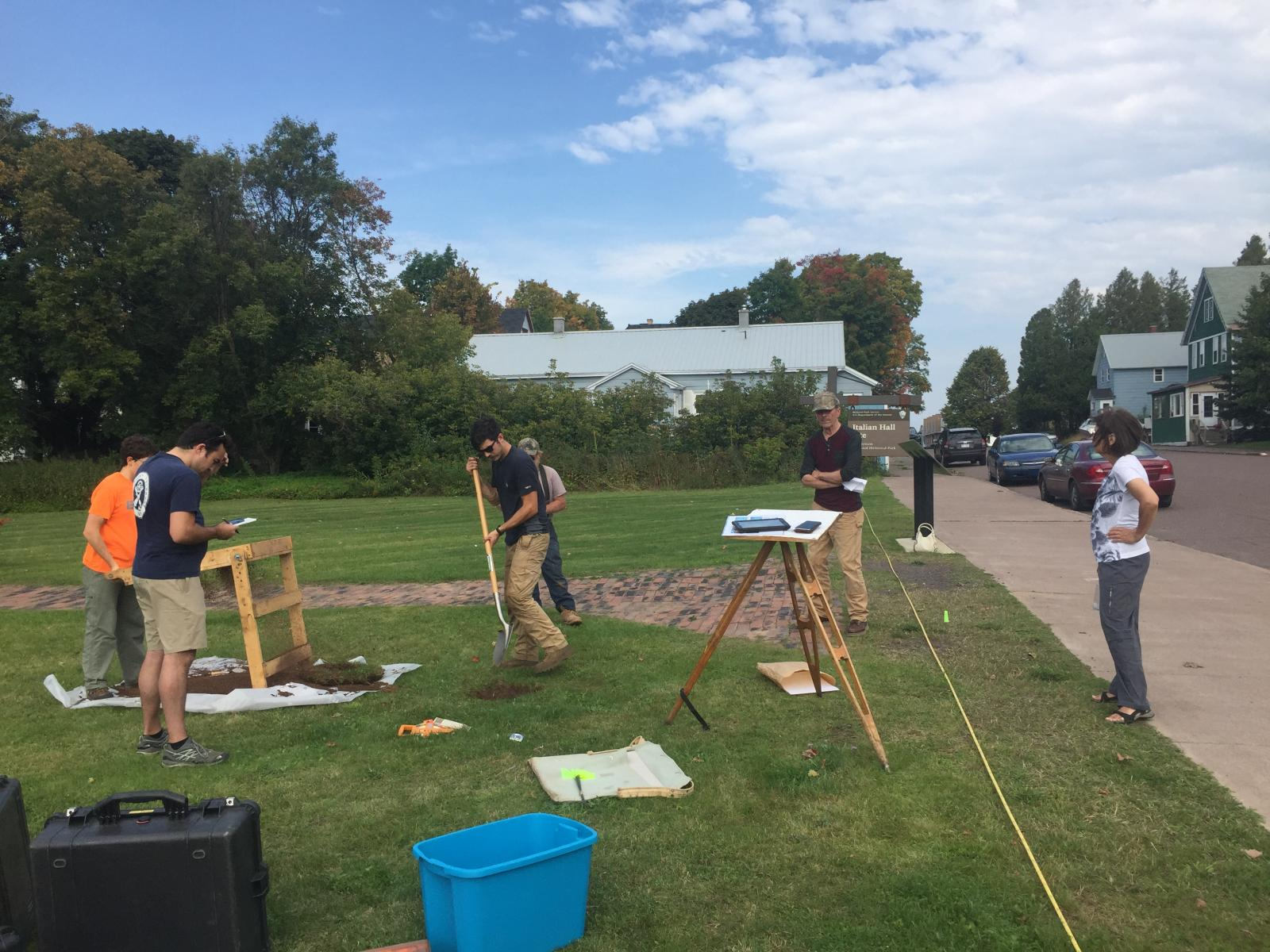 A group of 6 people working at an archaeological site, a grassy area, with brick walkway and sidewalk.