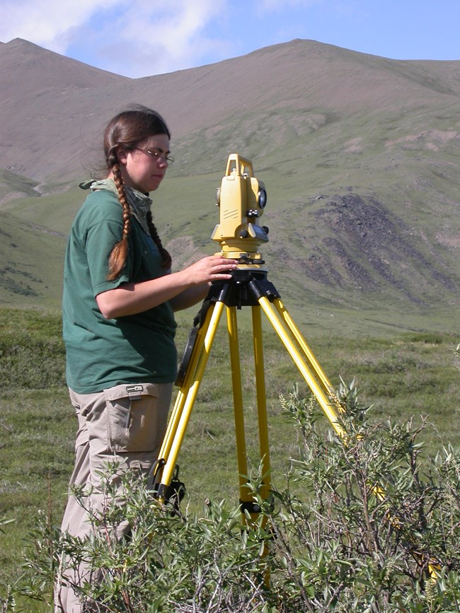 Archeologist conducts field work looking through a tool with mountains in the background.