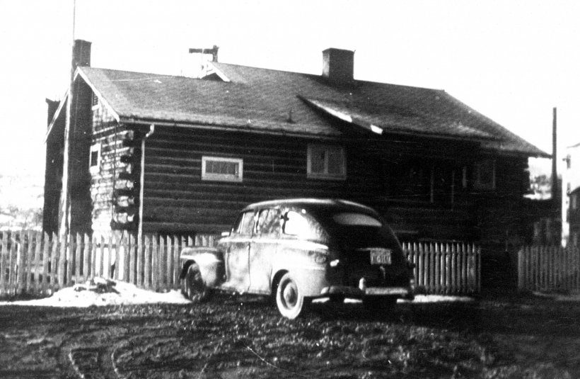 Historic log cabin with angled roof, two chimneys, classic car parked in front outside picket fence.