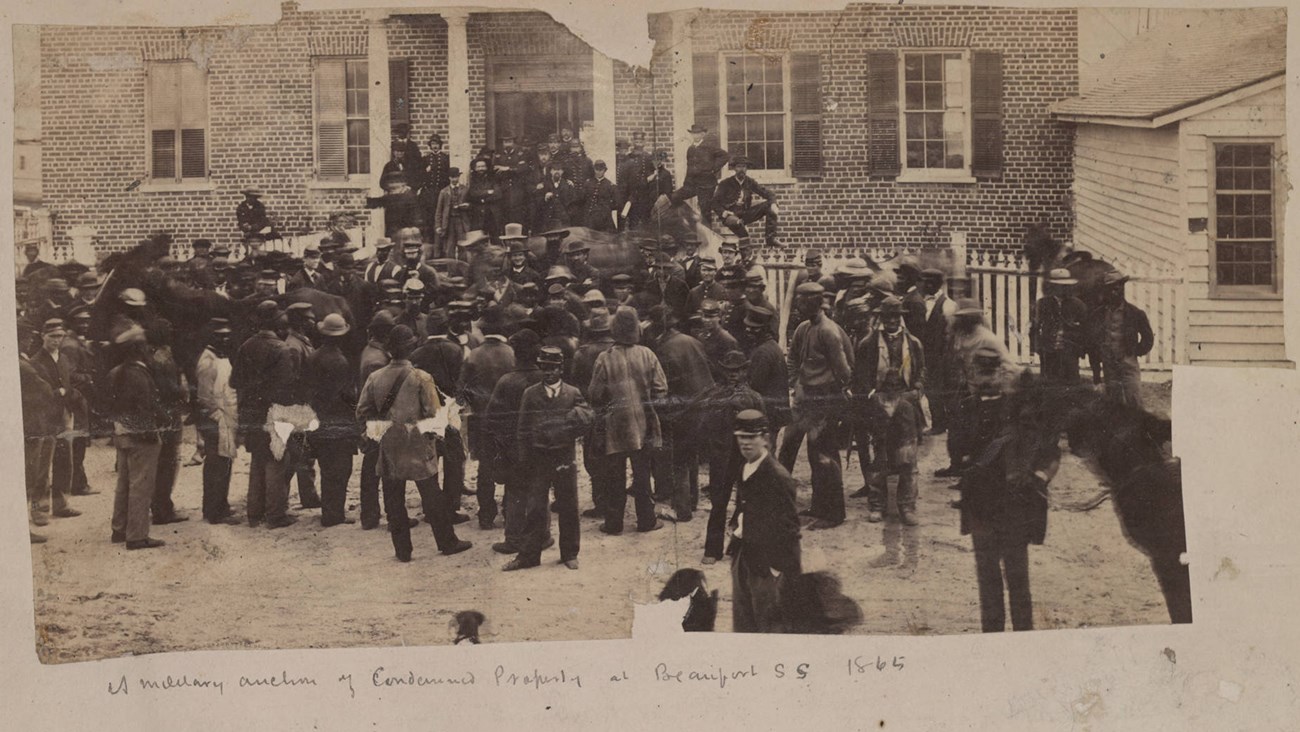 A large group of men stand in front of a building looking at an auctioneer.