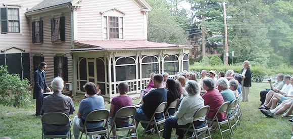 A group of people sit in chairs on the lawn of a Victorian-style house
