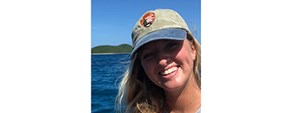 Smiling woman with blond hair blowing in the wind and an NPS ball cap in a boat with sunlit blue ocean and distant green shore behind her