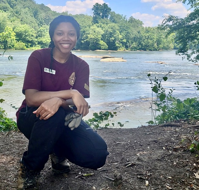 Intern Autumn Davis kneels in front of a a body of water with green trees behind it