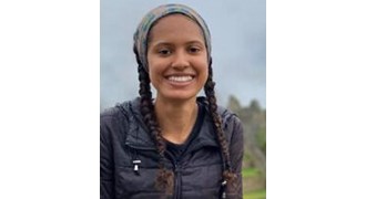 Fachon wearing a bandana, braids, and a black puffy vest, outside at a scenic vista smiling at the camera.