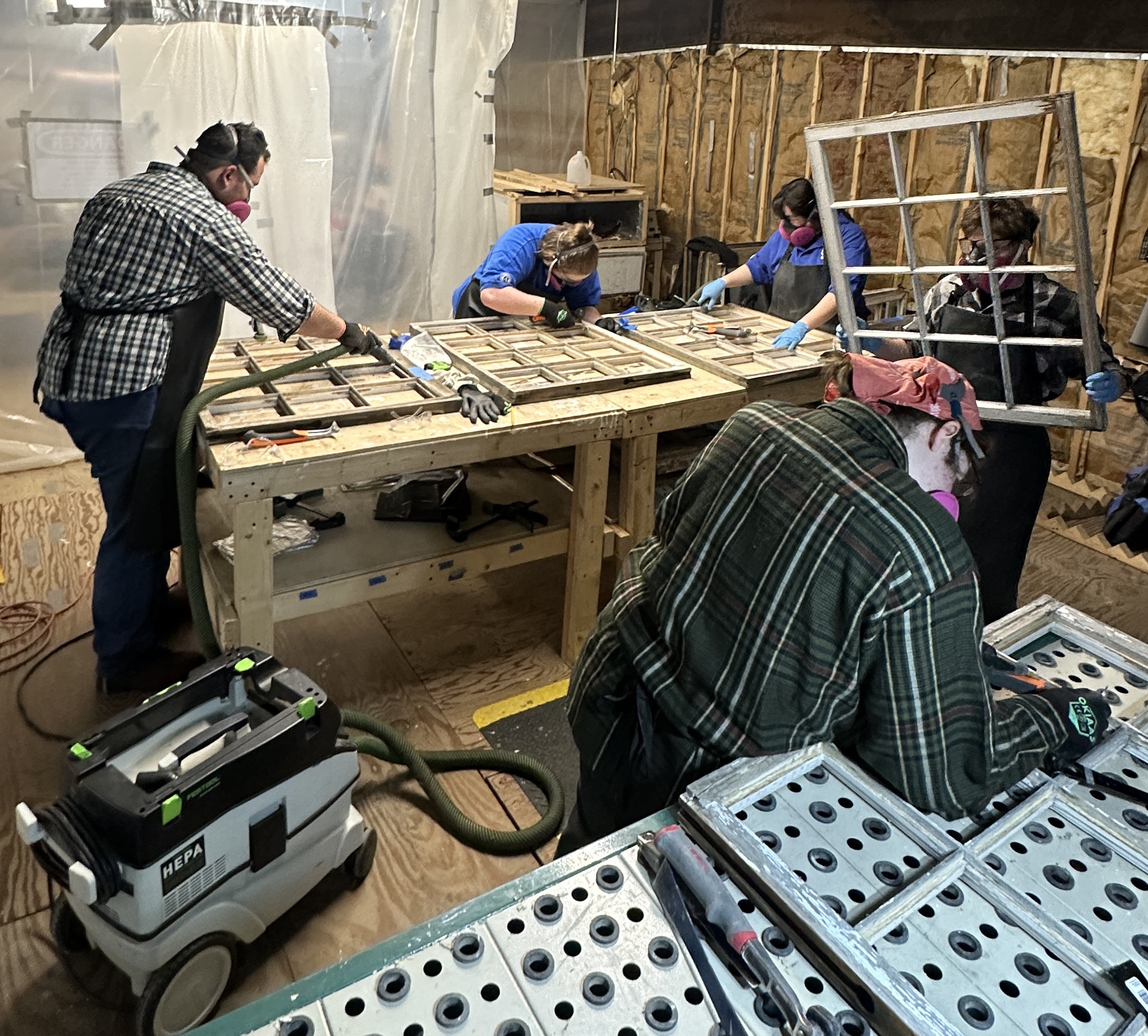 Five crew members working on windows. All of them are wearing protective gear such as respirators, gloves, safety glasses, and aprons, and using tools such as scrapers and vacuums.