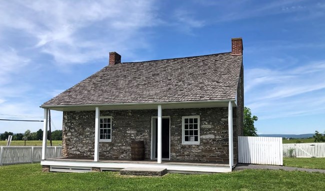 A one-story gray stone house with a covered porch and a wood-shingle roof with two brick chimneys sits on a green grass lawn enclosed by a low rectangular white picket fence. Trees and distant hills are visible in the background.