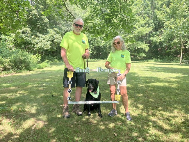 Two volunteers wearing bright green shirts holding a BARK Ranger photo frame. A black dog with a bright green bandana around his neck is sitting in the photo frame.