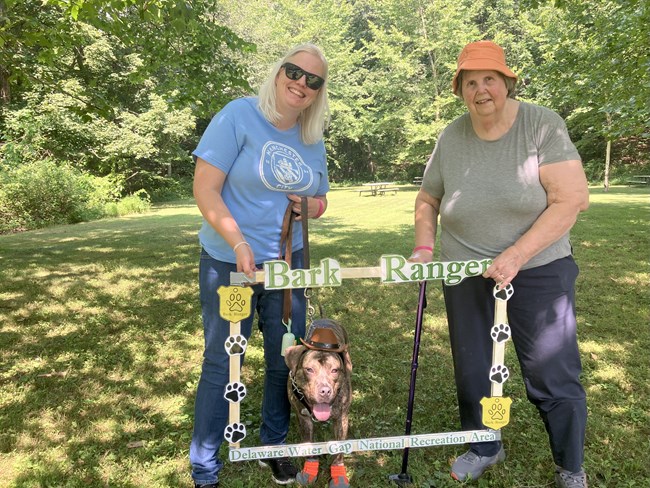 Two visitors hold a BARK Ranger photo frame around a dog.