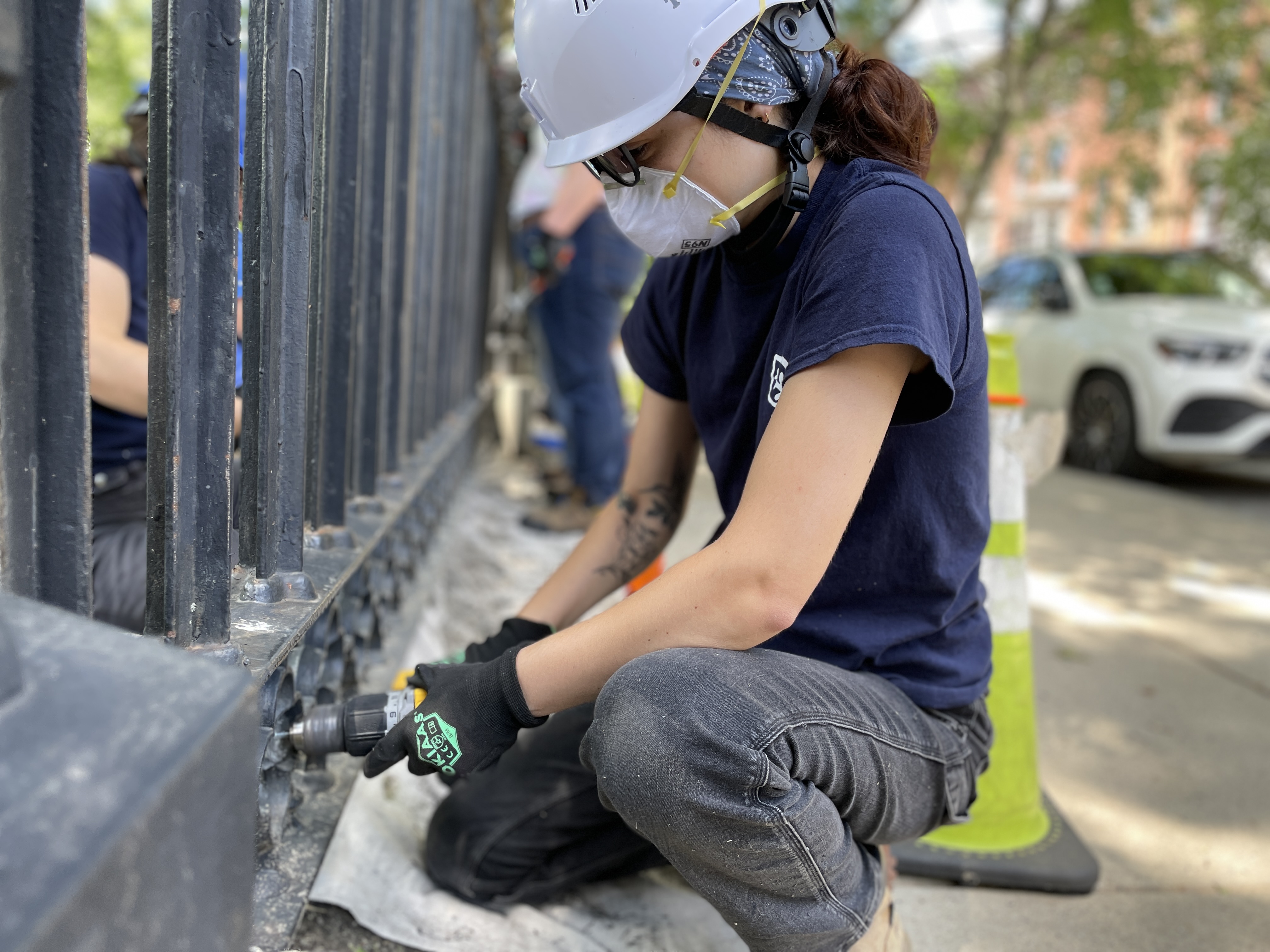 A crew member wearing a hard hat, gloves, and an N95 mask squatting at the bottom of a wrought-iron fence. The fence is dirty and has chipping paint in a few spots. The crew member is holding a drill with a wire brush attachment up to the fence.