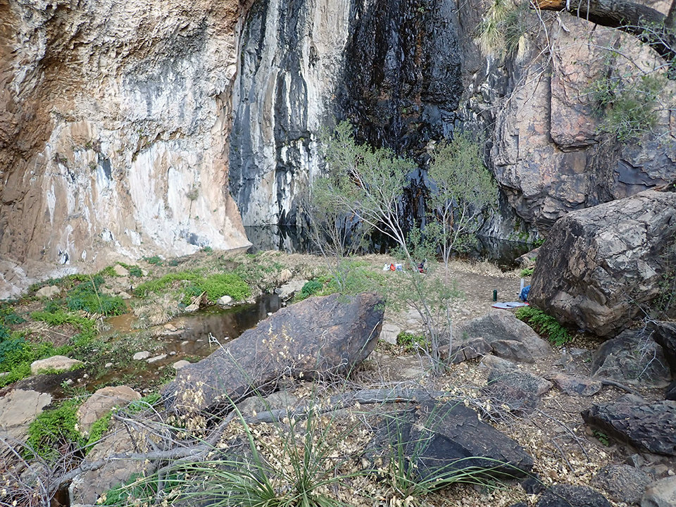 A large pool of water at the base of a steep rock wall where water flows into the pool and forms a stream surrounded by boulders and green vegetation.
